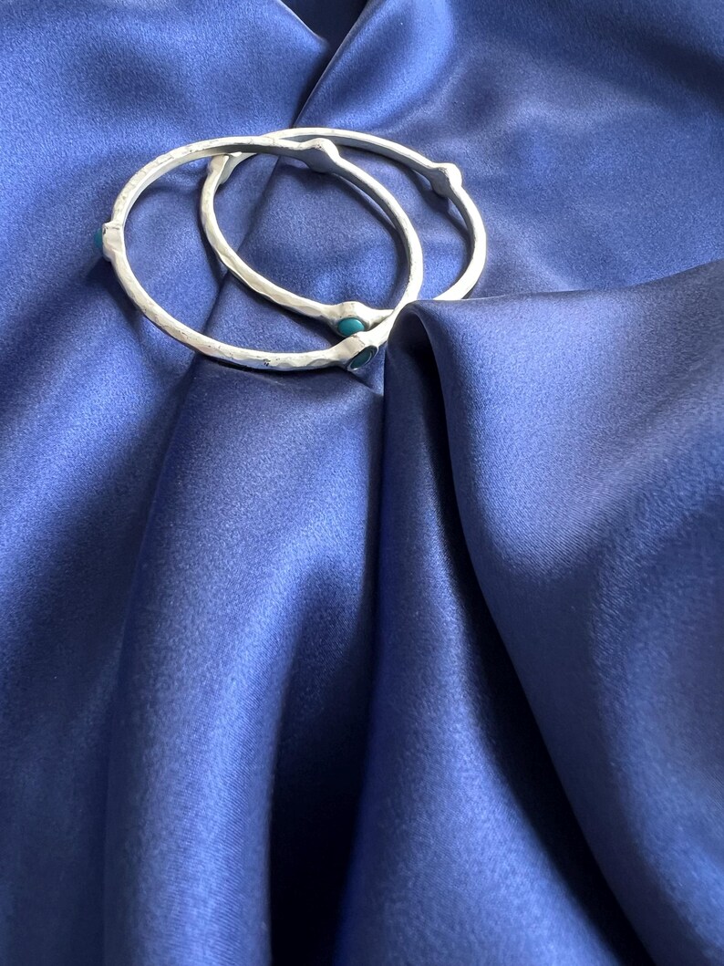 K&ouml;nnte beinhalten: Zwei silberne Armreifen mit t&uuml;rkisfarbenen Steinen, die in das Metall eingefasst sind. Die Armreifen liegen auf einem blauen Satin-Stoff.
