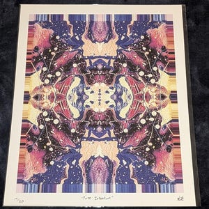 Puede incluir: Una impresión artística enmarcada con un diseño abstracto simétrico. La obra presenta una mezcla de colores, incluyendo rosa, morado, azul y amarillo, con motas blancas. El título "Pure Intention" está impreso en la parte inferior, junto con el número de edición 19/20.