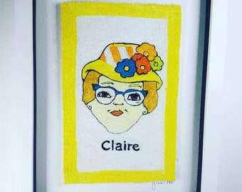 May include: Embroidered portrait of a woman with a yellow hat and flowers, wearing glasses, with the name "Claire" written below. The portrait is framed in a white frame with a yellow mat.