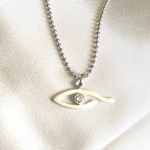 Könnte beinhalten: Halskette mit silberfarbener Kette und einem Anhänger in Augenform. Der Anhänger ist cremefarben mit einem kleinen, runden, klaren Stein in der Mitte. Die Kette besteht aus kleinen, runden Silberperlen.