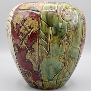 May include: A handcrafted ceramic vase with a unique design. The vase features sections of white, red, green, and gold colours. The green section has an embossed fish-like design. The vase has a textured surface and a rounded shape.
