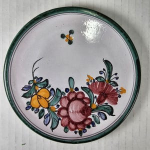 Peut inclure: Une assiette en céramique blanche avec un bord vert et un motif floral. L'assiette présente un motif peint à la main de fleurs rouges, jaunes et bleues avec des feuilles vertes. L'assiette est une pièce décorative.
