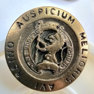 May include: Aged brass-toned circular emblem with the words "AUSPICIUM" and "MELIORIS" around the edge. The centre features a heraldic lion holding a flag. The words "REINO AVI" are also visible.