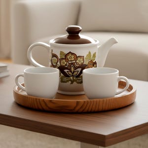 Peut inclure: Une théière blanche avec un couvercle marron et un motif floral repose sur un plateau en bois avec deux tasses blanches. La théière a une anse et un bec courbés. Le plateau et la table sont en bois.