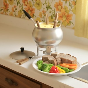 May include: A silver fondue pot with wooden skewers, a lid, and a plate of food. The plate contains bread cubes, broccoli florets, sliced carrots, red and yellow bell peppers, and green beans. The fondue pot sits on a metal stand.