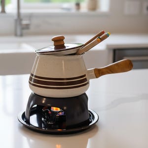 Peut inclure: Une fondue crémeuse avec des rayures marron et une poignée en bois est posée sur un brûleur noir avec une flamme visible. Deux fourchettes à fondue en bois reposent sur le pot. La pot a un couvercle avec un bouton en bois. Articles de cuisine.