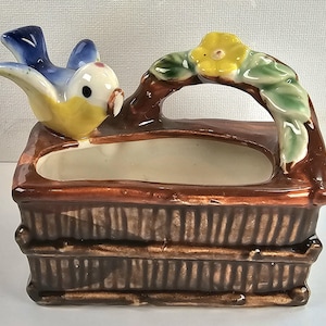 May include: A vintage ceramic basket planter with a blue and yellow bird on the side. The handle is decorated with a yellow flower and green leaves. The planter has a brown exterior and a cream interior. A charming decorative piece.