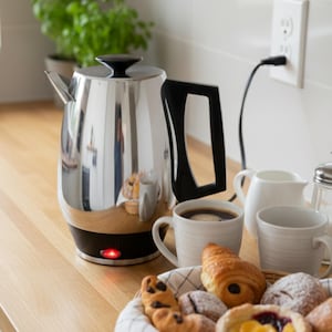 May include: A polished stainless steel electric kettle with a black handle and base on a wooden surface. Two mugs of coffee, pastries, and a basket are nearby, suggesting breakfast. A red light indicates the kettle is on.