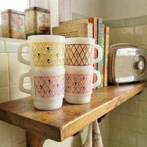 May include: Four white milk glass mugs with D-shaped handles, arranged in two stacks. Each mug features a repeating diamond lattice pattern with small floral accents in yellow, brown, pink, and orange colours printed on the exterior surface.