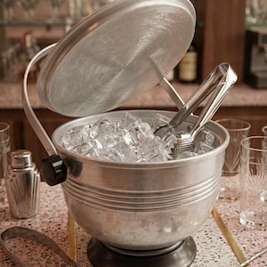 May include: A brushed metal ice bucket, filled with ice cubes, with a lid and handle. Ice tongs are inside the bucket. The scene suggests a bar setting, with a cocktail shaker and bar spoons also visible, perfect for making cocktails.