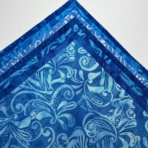 May include: A stack of four folded fabric squares arranged in a triangular formation. Each layer features a distinct blue batik pattern, ranging from deep navy to light cyan, with intricate floral and swirling motifs printed on the cotton textile.