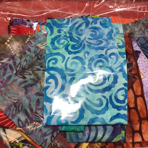 May include: A clear plastic bag filled with colorful fabric scraps. The fabrics feature various patterns, including swirls, floral designs, and abstract shapes in shades of blue, green, orange, and purple. The bag is sealed with a white zipper.