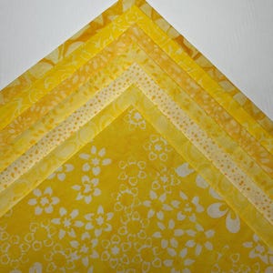 Puede incluir: Una pila de cuadrados de tela amarillos con varios diseños florales y estampados. Las telas varían en tonos de amarillo, de claro a dorado, y están dispuestas en una composición en capas y en ángulo. Adecuado para acolchar o manualidades.