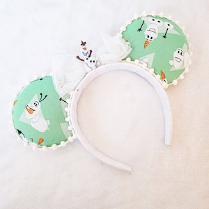 May include: A headband featuring two round ears covered in light green fabric with a repeating Olaf character print. The ears are trimmed with small white pom-poms. A white sheer bow sits between the ears, accented with a small Olaf figure.