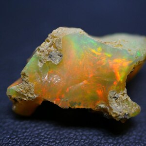 Puede incluir: Una piedra preciosa de ópalo en bruto con una mezcla de colores, incluyendo naranja, verde y amarillo. La piedra tiene una superficie texturizada e irregular con una apariencia natural y terrosa. El ópalo se encuentra sobre un fondo oscuro.