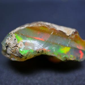 Puede incluir: Una piedra preciosa de ópalo en bruto con una mezcla de colores, incluyendo verde, rojo y amarillo. El ópalo tiene una apariencia translúcida con un exterior rugoso y texturizado. La gema se encuentra sobre un fondo oscuro.