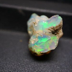 Può includere: Una gemma di opale grezza con una vibrante esposizione di colore. La pietra mostra lampi di verde, blu e giallo, su una base chiara. L'opale ha un aspetto naturale e non lucidato.