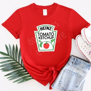 May include: Red t-shirt with a white Heinz Tomato Ketchup logo printed on the front. The shirt is tied in a knot at the waist. A pair of white sneakers and a pair of light blue denim shorts are also in the image.