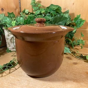 May include: A brown ceramic pot with a lid, suitable for cooking or storage. The pot has a rounded shape with a slightly flared rim and a small knob on the lid. The pot is approximately 7 inches in diameter.
