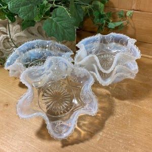 May include: Three opalescent glass bowls with ruffled edges and starburst patterns. The bowls have a delicate, translucent appearance and are arranged on a wooden surface. The bowls are likely vintage or antique decorative items.