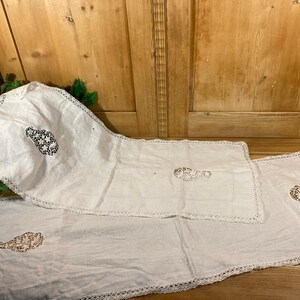 May include: White linen table runners and placemats with decorative lace edges and embroidered floral designs. The set includes a long runner, a shorter runner, and a square placemat. The embroidery features black and tan colors.