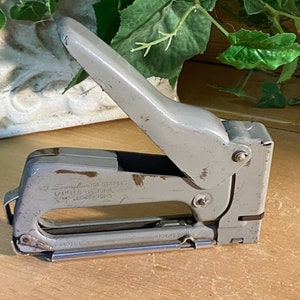 May include: A gray metal staple gun with a silver staple remover. The gun has a handle and a lever to operate it. The text on the gun reads "Use Swingline 101 Staples" and "Varieties No. 101-4, 101-5, 101-6". The gun is labeled "Empty".