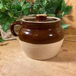 May include: A round ceramic crock featuring a two-tone design with a glossy dark brown glazed upper section and a matte beige lower section. The pot includes a matching lid with a small knob handle and a single curved side handle for gripping.