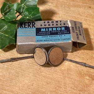 May include: Vintage Kerr Mirror set with two small, round mirrors and a box. The box is labeled "Kerr Mirror" with text about the rhodium-coated lens and cone socket. The mirrors have metal handles and are on a wooden surface.