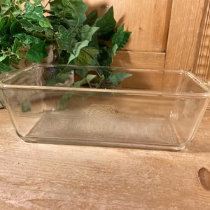 May include: A clear rectangular glass loaf pan featuring a decorative embossed pattern on the side. The design includes horizontal lines and a central scrollwork motif. The glass is transparent with a smooth finish and integrated handles on the ends.