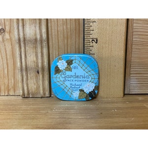 May include: A blue and gold compact with a white and green floral design. The text on the compact reads "Gardenia Face Powder Richard Hudnut New York Paris".