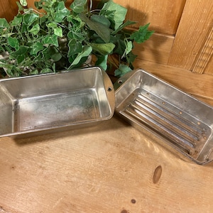 May include: Two rectangular metal loaf pans with metallic silver finishes. One pan features a smooth interior base, while the other pan has a ribbed bottom surface with small drainage holes. Both pans have small handles on each end with holes.