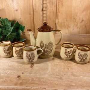 May include: A ceramic tea set featuring a teapot with a brown lid and four matching mugs. Each piece is cream-colored with a speckled finish and decorated with brown line-art illustrations of roosters and hens, accented by brown rims.
