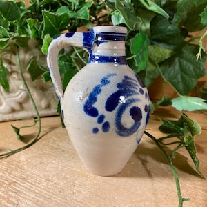 May include: A small, off-white ceramic pitcher featuring a single handle and a narrow neck. The vessel is decorated with hand-painted cobalt blue floral and swirling patterns, with horizontal blue bands encircling the rim and the base of the neck.