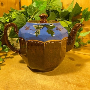May include: A brown teapot with a blue and gold floral design. The teapot has a rounded body and a spout on the side. The teapot is sitting on a wooden surface.