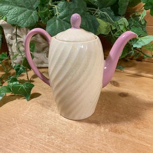 May include: Cream-colored ceramic teapot with a spiral design, featuring a pink handle, spout, and lid knob. The teapot is set against a wooden surface with green foliage in the background. A decorative item for serving tea.