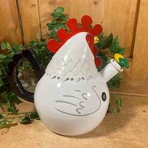 May include: A white ceramic rooster-shaped teapot with a red comb and yellow beak. The teapot has a black handle and a green and yellow spout. The design includes gray lines to represent feathers. The teapot is surrounded by green foliage.