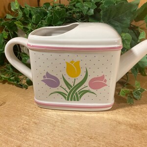 May include: White ceramic watering can with a pink trim and a floral design featuring yellow, purple, and pink tulips. The watering can has a handle and a spout, and is decorated with small gray polka dots.