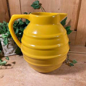 May include: A bright yellow ceramic pitcher with a rounded, bulbous body featuring horizontal ribbed detailing. The vessel has a wide, curved handle on the left side and a small, integrated pouring spout at the top rim, finished in a smooth glaze.