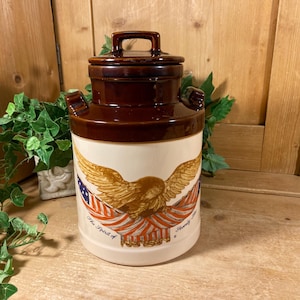 May include: A cylindrical ceramic crock with a dark brown lid and upper rim, featuring a cream-colored base. The front displays a golden eagle graphic perched above a draped American flag with the text The Spirit of Seventy-Six printed below.