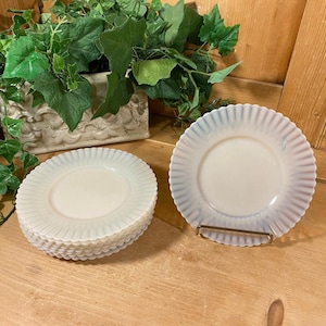 May include: A set of six white milk glass plates with scalloped edges. The plates are stacked on top of each other and have a light blue iridescent sheen.