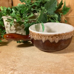 May include: A brown ceramic soup bowl with a handle. The bowl has a cream-colored interior and a brown exterior with a lighter brown drip glaze. The handle is a dark brown color.