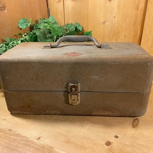 May include: A weathered, rectangular metal toolbox with a hinged lid and a sturdy handle. The toolbox is a muted brown color with a tarnished appearance, suggesting age and use. A small, red and white emblem is visible on the top. The toolbox is closed with a metal clasp.