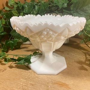 May include: A white, vintage-style pedestal bowl with a scalloped rim and embossed diamond and floral patterns. The bowl sits on a hexagonal base. Green foliage is in the background.
