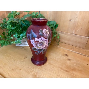May include: A burgundy glass vase with a floral design painted on the front. The flowers are pink and white with green leaves.