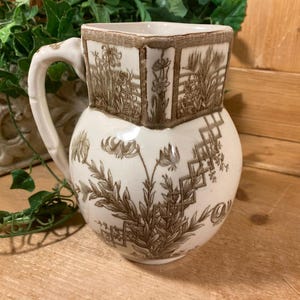 May include: Cream-colored ceramic pitcher with a square top and rounded body. The pitcher features a brown floral and geometric pattern. It has a curved handle and is placed on a wooden surface.