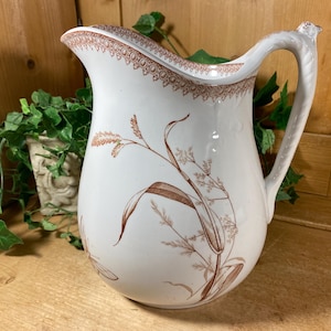 May include: White ceramic pitcher with a brown wheat stalk design. The pitcher has a decorative brown border around the rim and a curved handle. The pitcher is a vintage style and is suitable for serving water or other beverages.
