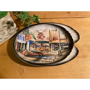 May include: A white ceramic ashtray with a painted scene of a Parisian street with a windmill, buildings, and people. The text "Paris, Le Moulin Rouge" is written on the bottom of the ashtray.