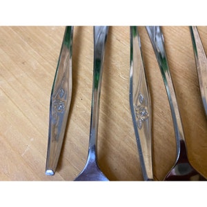 May include: Five silver spoons with a rose design on the handle. The spoons are arranged in a row on a wooden surface.