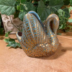 May include: A gold and teal ceramic swan figurine. The swan has a textured, cracked pattern design. The interior of the swan is teal. The swan is sitting on a wooden surface with greenery in the background.