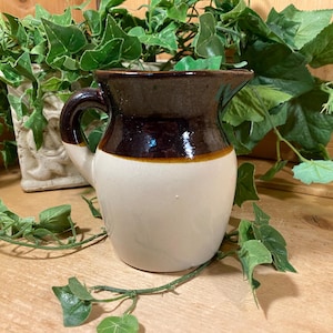 May include: A ceramic pitcher featuring a two-tone design with a glossy dark brown glaze on the upper portion and a smooth cream-colored base. The vessel includes a curved handle on the side and a small pouring spout at the top rim.
